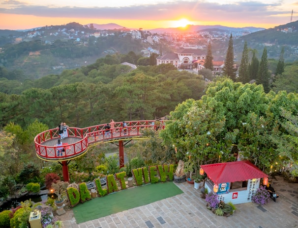 A scenic viewpoint overlooking a lush green valley with a vibrant sunset in the background. A circular viewing platform with a red railing is situated among the trees, where a few people are enjoying the view. Below, there is a small structure with a red roof, adorned with signs and lanterns. The foreground features a neatly arranged garden with letters spelling out 'DA LAT VIEW'. Houses are scattered on the distant hills, surrounded by dense forest.