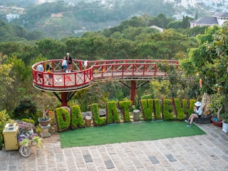 A scenic location features a wooden observation deck with a red railing elevated above lush greenery. Several people enjoy the view from the deck. In front of the deck, large letters made from plants spell out 'DA LAT VIEW.' A woman in a hat sits on a stone bench nearby, while potted plants and a vintage bicycle adorn the area.