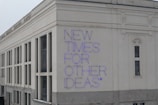 A large building facade with a modern architectural style, featuring white and beige exterior walls. The phrase 'NEW TIMES FOR OTHER IDEAS' is prominently displayed in purple neon letters, creating a bold contrast against the light background.