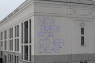 A large building facade with a modern architectural style, featuring white and beige exterior walls. The phrase 'NEW TIMES FOR OTHER IDEAS' is prominently displayed in purple neon letters, creating a bold contrast against the light background.