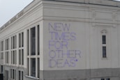 A large building facade with a modern architectural style, featuring white and beige exterior walls. The phrase 'NEW TIMES FOR OTHER IDEAS' is prominently displayed in purple neon letters, creating a bold contrast against the light background.