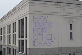 A large building facade with a modern architectural style, featuring white and beige exterior walls. The phrase 'NEW TIMES FOR OTHER IDEAS' is prominently displayed in purple neon letters, creating a bold contrast against the light background.