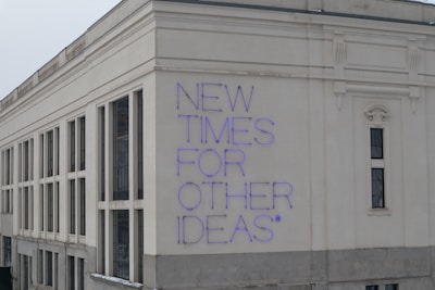 A large building facade with a modern architectural style, featuring white and beige exterior walls. The phrase 'NEW TIMES FOR OTHER IDEAS' is prominently displayed in purple neon letters, creating a bold contrast against the light background.