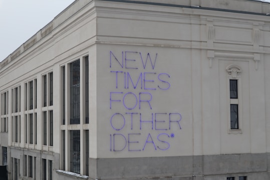 A large building facade with a modern architectural style, featuring white and beige exterior walls. The phrase 'NEW TIMES FOR OTHER IDEAS' is prominently displayed in purple neon letters, creating a bold contrast against the light background.
