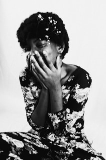 A person is captured in a black and white photograph, seemingly caught in a moment of joy or laughter, with their hands partially covering their face. They are wearing a floral-patterned outfit and have small flowers adorning their hair.