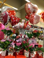 A vibrant display of Valentine's Day-themed decorations featuring an assortment of heart-shaped balloons with romantic messages, surrounded by a variety of potted flowers in colorful containers. The scene is adorned with red and pink hues, emphasizing the festive and romantic atmosphere.