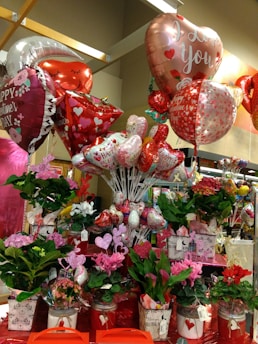 A vibrant display of Valentine's Day-themed decorations featuring an assortment of heart-shaped balloons with romantic messages, surrounded by a variety of potted flowers in colorful containers. The scene is adorned with red and pink hues, emphasizing the festive and romantic atmosphere.