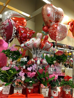 A vibrant display of Valentine's Day-themed decorations featuring an assortment of heart-shaped balloons with romantic messages, surrounded by a variety of potted flowers in colorful containers. The scene is adorned with red and pink hues, emphasizing the festive and romantic atmosphere.