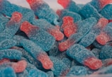 A close-up of colorful gummy candies shaped like bottles, covered in sugar. These candies have a gradient of blue and red colors with a textured sugary coating.