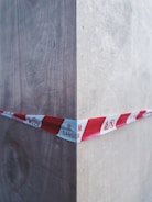 A red and white caution tape marked with 'DANGER' in multiple languages stretches across a wooden corner, indicating a hazardous area. The wood appears to be unfinished and shows grain texture, with shadows and highlights accentuating the corner.