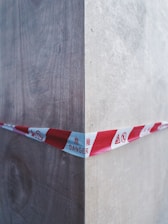 A red and white caution tape marked with 'DANGER' in multiple languages stretches across a wooden corner, indicating a hazardous area. The wood appears to be unfinished and shows grain texture, with shadows and highlights accentuating the corner.