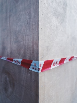 A red and white caution tape marked with 'DANGER' in multiple languages stretches across a wooden corner, indicating a hazardous area. The wood appears to be unfinished and shows grain texture, with shadows and highlights accentuating the corner.