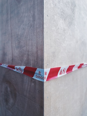 A red and white caution tape marked with 'DANGER' in multiple languages stretches across a wooden corner, indicating a hazardous area. The wood appears to be unfinished and shows grain texture, with shadows and highlights accentuating the corner.