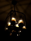 A dimly lit chandelier containing several glowing candles is encased in a transparent, geometric glass frame. The dark background accentuates the soft, warm glow of the candlelight, creating intricate shadow patterns.