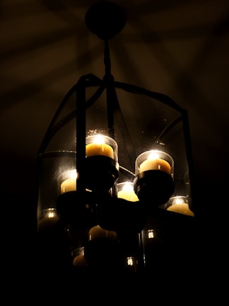 A dimly lit chandelier containing several glowing candles is encased in a transparent, geometric glass frame. The dark background accentuates the soft, warm glow of the candlelight, creating intricate shadow patterns.