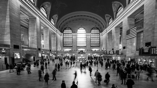 A grand hall with high arched windows and ornate ceilings, bustling with a crowd of people. The space is expansive, illuminated by natural light filtering through the large windows and chandeliers. An American flag hangs prominently, and various information boards are visible throughout.