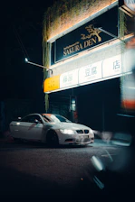 A white car with its headlights on is parked in front of a restaurant called Sakura Den, which has a brightly lit sign with Japanese characters. The building has greenery hanging from the top, and the scene is set at night with a moody atmospheric light.