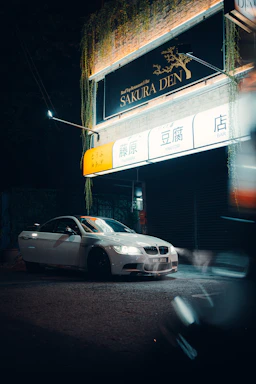 A white car with its headlights on is parked in front of a restaurant called Sakura Den, which has a brightly lit sign with Japanese characters. The building has greenery hanging from the top, and the scene is set at night with a moody atmospheric light.