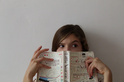 A person is holding a book titled 'Little Women' in front of their face with only their eyes visible. They appear to be looking to the side. The book cover is decorated with illustrations and text.
