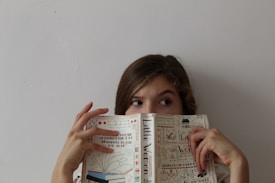 A person is holding a book titled 'Little Women' in front of their face with only their eyes visible. They appear to be looking to the side. The book cover is decorated with illustrations and text.