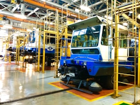 A spacious industrial workshop with multiple blue and white rail maintenance vehicles positioned on tracks. The vehicles are surrounded by a structural framework of yellow metal platforms and ladders, used for maintenance purposes. Overhead cranes and a lattice of beams and lighting fixtures dominate the ceiling, providing a busy industrial atmosphere.