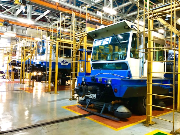 A spacious industrial workshop with multiple blue and white rail maintenance vehicles positioned on tracks. The vehicles are surrounded by a structural framework of yellow metal platforms and ladders, used for maintenance purposes. Overhead cranes and a lattice of beams and lighting fixtures dominate the ceiling, providing a busy industrial atmosphere.