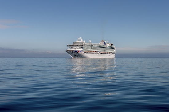 A large cruise ship is sailing on calm, open waters under a clear blue sky. The ship is predominantly white with some colorful markings and has multiple decks with various structures visible on top.