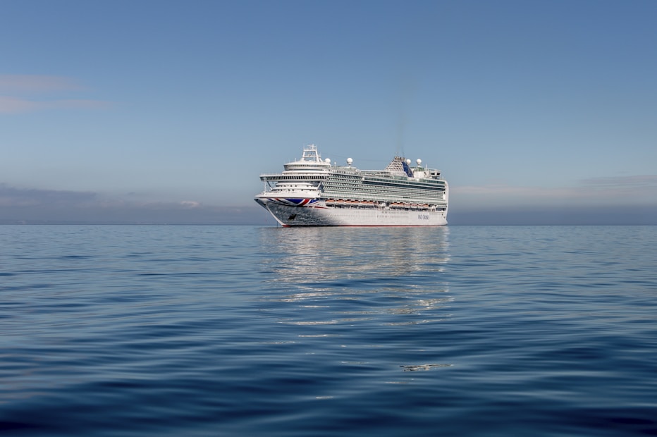A large cruise ship is sailing on calm, open waters under a clear blue sky. The ship is predominantly white with some colorful markings and has multiple decks with various structures visible on top.
