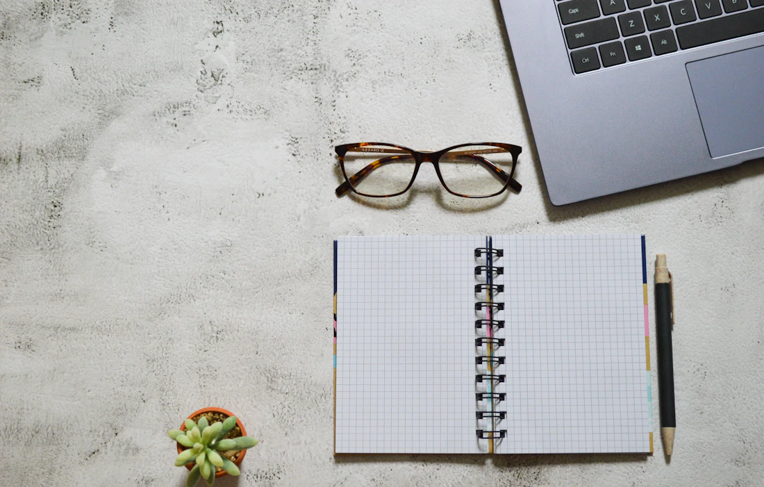A notebook, reading glasses and a laptop laid out on a light desk