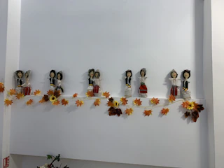 A line of small, handcrafted dolls dressed in traditional attire is displayed on a shelf. They are accompanied by an autumn-themed garland of orange and yellow leaves.