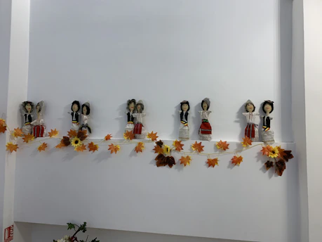 A line of small, handcrafted dolls dressed in traditional attire is displayed on a shelf. They are accompanied by an autumn-themed garland of orange and yellow leaves.