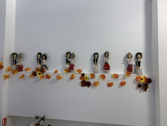 A line of small, handcrafted dolls dressed in traditional attire is displayed on a shelf. They are accompanied by an autumn-themed garland of orange and yellow leaves.