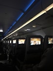 An interior view of an airplane cabin featuring several rows of seats. Each seat is equipped with a personal screen displaying a welcome message and an image of rock formations at sunset. The lighting is dim with a blue ambiance from the overhead compartments.
