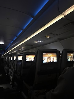 An interior view of an airplane cabin featuring several rows of seats. Each seat is equipped with a personal screen displaying a welcome message and an image of rock formations at sunset. The lighting is dim with a blue ambiance from the overhead compartments.