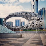 A large, futuristic building featuring a torus-shaped structure covered in Arabic calligraphy is set against a backdrop of modern skyscrapers. The scene is expansive with a clear sky and a tiled plaza in the foreground, which highlights the architectural design.