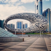 A large, futuristic building featuring a torus-shaped structure covered in Arabic calligraphy is set against a backdrop of modern skyscrapers. The scene is expansive with a clear sky and a tiled plaza in the foreground, which highlights the architectural design.