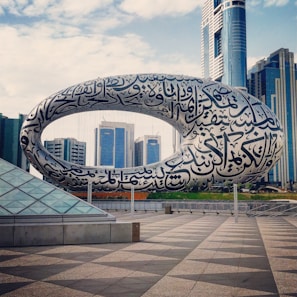 A large, futuristic building featuring a torus-shaped structure covered in Arabic calligraphy is set against a backdrop of modern skyscrapers. The scene is expansive with a clear sky and a tiled plaza in the foreground, which highlights the architectural design.