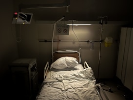 A hospital bed with an intravenous stand and medical equipment surrounding it. The bed is unoccupied with crumpled white sheets and a pillow. There is a monitoring screen to the left, and a sign above the bed displaying the word 'NÜCHTERN' with other symbols. The lighting is dim and a sterile, clinical environment is suggested.