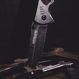 A close-up of a metal knife with an engraved logo on its blade, positioned vertically among several tools or pieces of metal on a dark surface. The lighting emphasizes the texture and details of the knife.