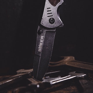 A close-up of a metal knife with an engraved logo on its blade, positioned vertically among several tools or pieces of metal on a dark surface. The lighting emphasizes the texture and details of the knife.