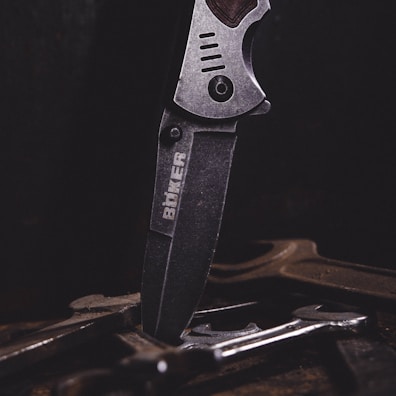 A close-up of a metal knife with an engraved logo on its blade, positioned vertically among several tools or pieces of metal on a dark surface. The lighting emphasizes the texture and details of the knife.