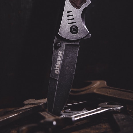 A close-up of a metal knife with an engraved logo on its blade, positioned vertically among several tools or pieces of metal on a dark surface. The lighting emphasizes the texture and details of the knife.