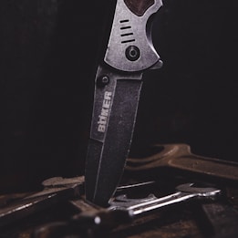 A close-up of a metal knife with an engraved logo on its blade, positioned vertically among several tools or pieces of metal on a dark surface. The lighting emphasizes the texture and details of the knife.