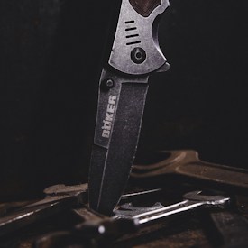 A close-up of a metal knife with an engraved logo on its blade, positioned vertically among several tools or pieces of metal on a dark surface. The lighting emphasizes the texture and details of the knife.