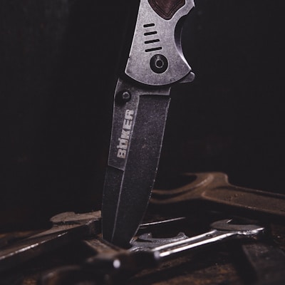 A close-up of a metal knife with an engraved logo on its blade, positioned vertically among several tools or pieces of metal on a dark surface. The lighting emphasizes the texture and details of the knife.