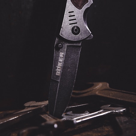 A close-up of a metal knife with an engraved logo on its blade, positioned vertically among several tools or pieces of metal on a dark surface. The lighting emphasizes the texture and details of the knife.