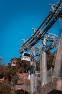 A cable car system with several gondolas suspended from steel cables, situated against a clear blue sky. The structure includes tall metal support towers and overhead wires. In the background, there are trees and a small building visible.