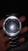 A shiny metallic wristwatch is being held in a hand. The watch face reads 'FIRE COLLECTION' and has a reflective surface. The metal band is composed of linked segments.