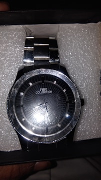 A sleek, metallic wristwatch with a reflective black dial and silver accents. The watch is branded with 'FIRE COLLECTION' and features a metal wristband. It is set against a textured white background within a box.