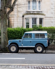 A vintage blue Land Rover is parked on the street in front of a stone building with large windows and neatly trimmed hedges. A large tree stands to the left of the vehicle, and the road is lined with cobblestones and asphalt.